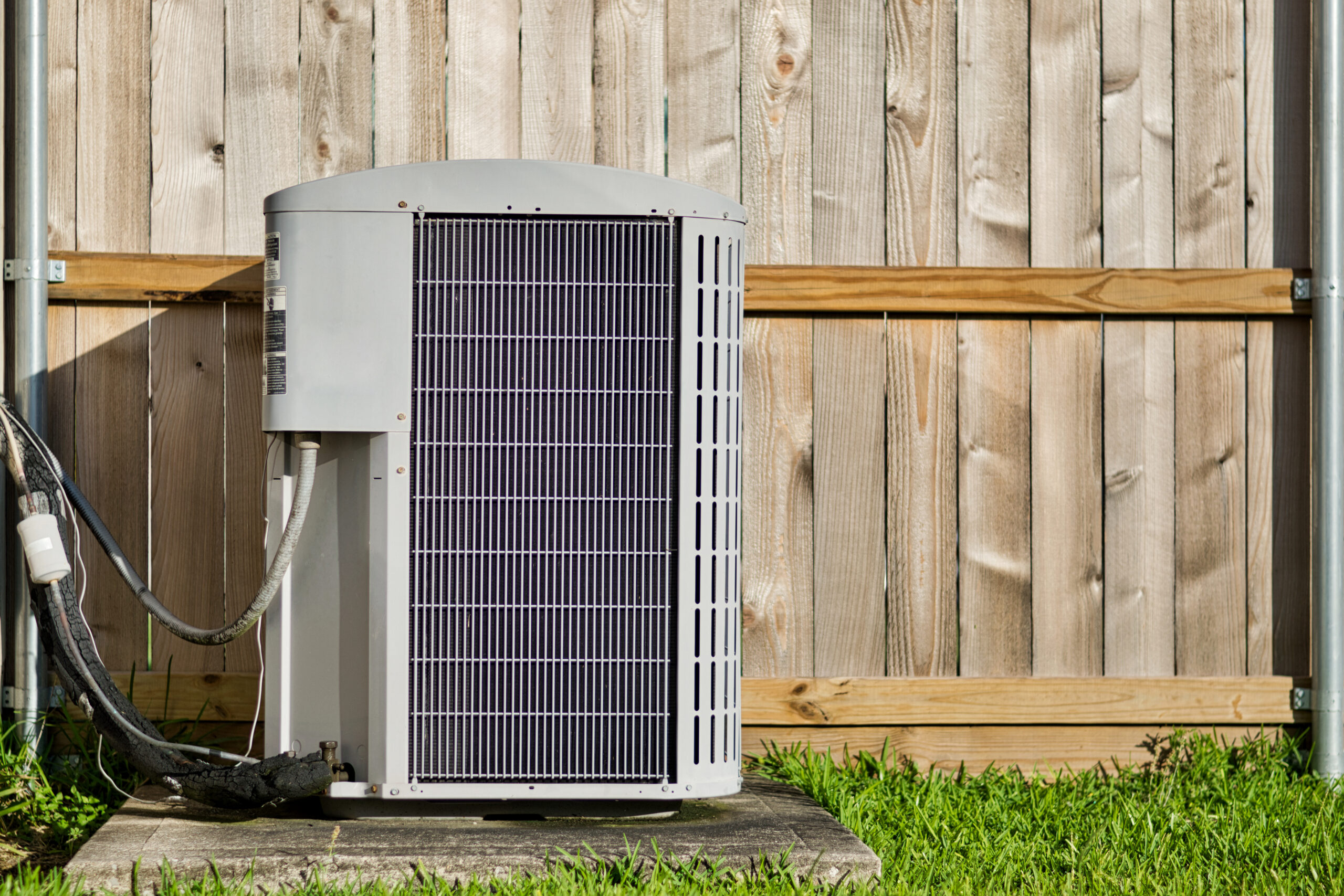 Outdoor AC condenser unit newly installed on a concrete pad against a wood fence in a residential backyard